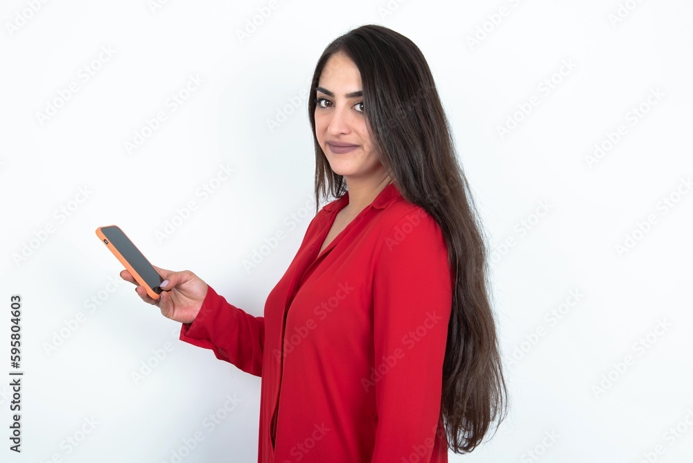 Rear view photo portrait of Young brunette woman wearing red shirt over white studio background using smartphone smiling Rear view photo portrait of Young brunette woman wearing red shirt over white studio background using smartphone smiling