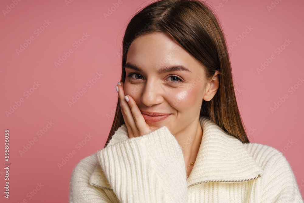Woman smiling and touching her cheek while standing isolated over pink wall