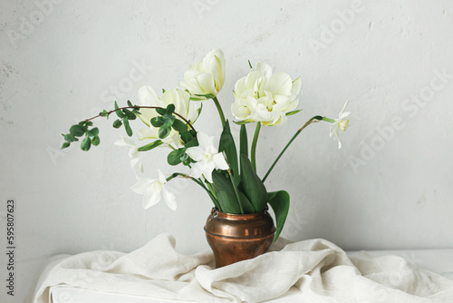 Beautiful white tulips and daffodils in vintage vase on wooden table against rustic wall. Happy Mothers day. Stylish simple spring bouquet, floral still life. Womens day. Space for text