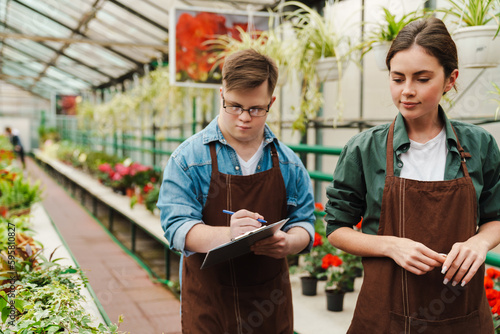 Man with down syndrome writing down notes while woman helping him to handle with flowers in greenhouse