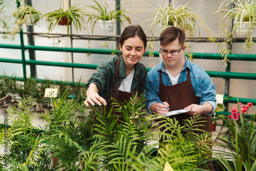Man with down syndrome writing down notes while woman helping him to handle with flowers in greenhouse photo