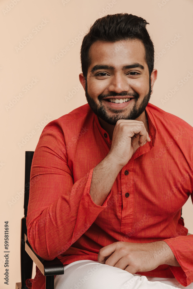 Young indian man smiling while sitting on chair isolated over beige background Young indian man smiling while sitting on chair isolated over beige background