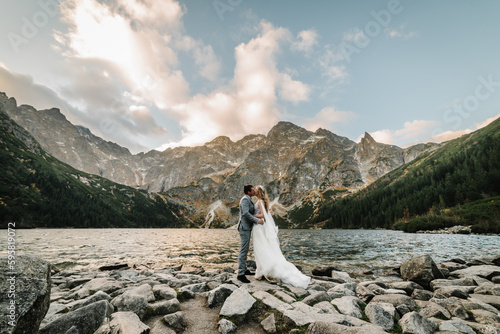 Romantic wedding couple in love standing on the stony shore of the Sea Eye lake in Poland. Scenic mountain view. The bride and groom. Morskie Oko. Tatra mountains.