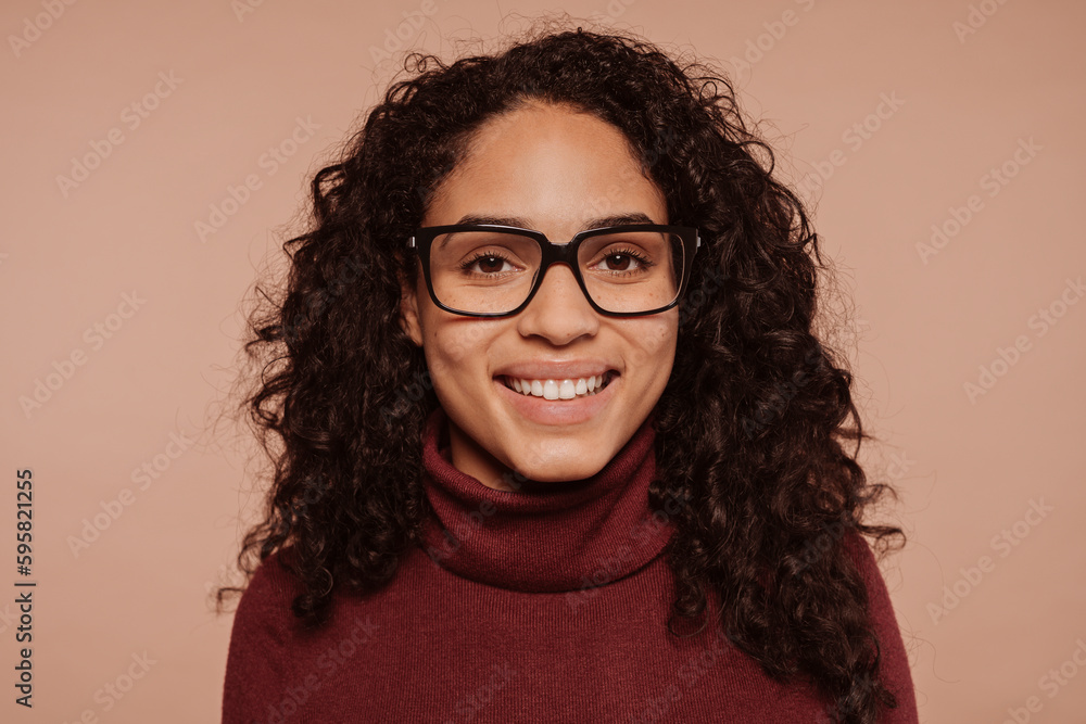 Close-up portrait of a happyy smiling multiracial brunette curly haired girl wears garnet turtleneck sweater and eyeglasses, at studio isolated over beige background.
