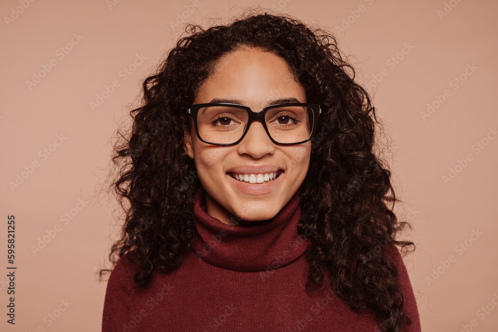 Close-up portrait of a happyy smiling multiracial brunette curly haired girl wears garnet turtleneck sweater and eyeglasses, at studio isolated over beige background. Close-up portrait of a happyy smiling multiracial brunette curly haired girl wears garnet turtleneck sweater and eyeglasses, at studio isolated over beige background.
