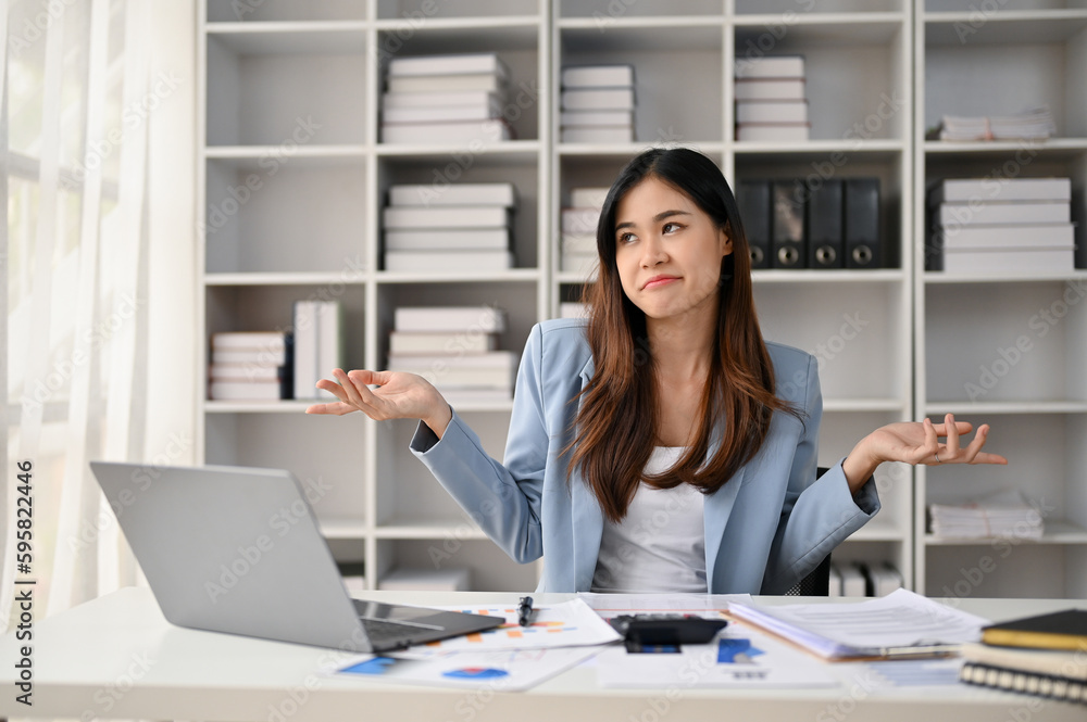 Confident Asian businesswoman sits at her office desk, smiling and shrugging her shoulders.