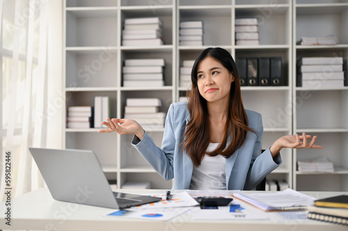 Fotografie Confident Asian businesswoman sits at her office desk, smiling and shrugging her shoulders