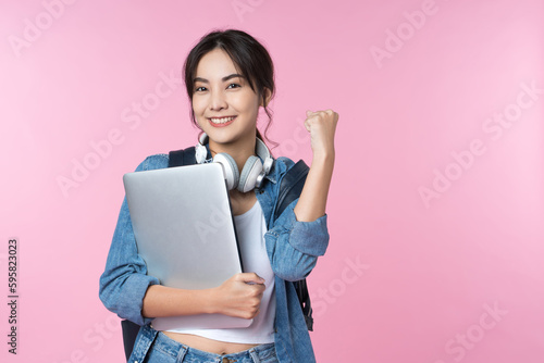 Portrait of smiling young Asian college student with laptop and backpack isolated over pink background.Cheer up success celebration concept photo