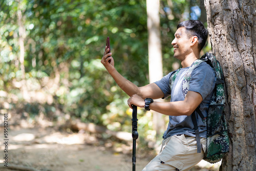 Handsome asian young male traveler with backpack, holding walking stick and hiking stick, standing in the forest with happiness, freedom in summer vacation, travel concept.