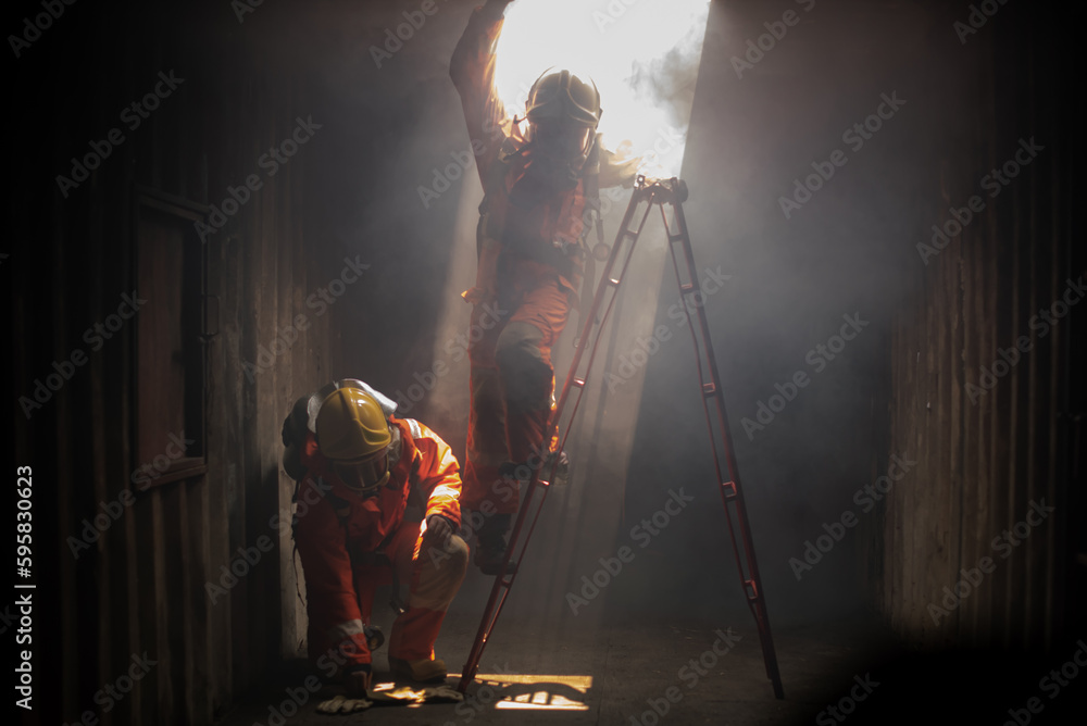 One firefighters checks the safety of the area while another attempts to climb a ladder in the galvanized room with soft light. One firefighters checks the safety of the area while another attempts to climb a ladder in the galvanized room with soft light.