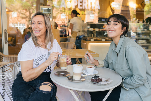 Smiling mother and daughter sitting on the terrace of a cafe having breakfast looking away