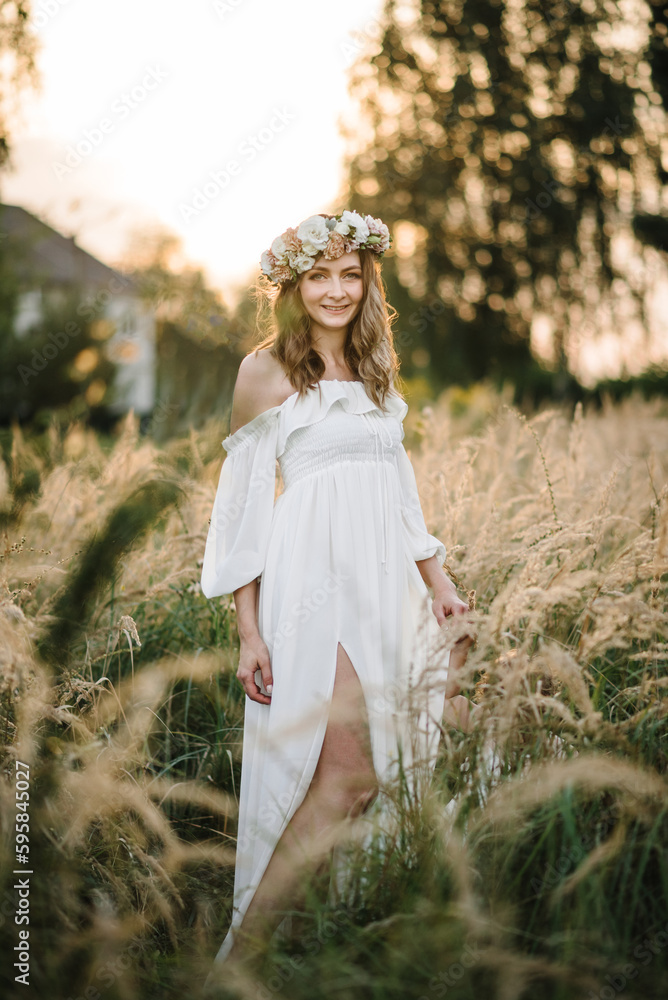 Naklejka premium A beautiful tender young girl in a white dress with a wreath of flowers on her head is standing in a spring field at sunset. Happy bride with blonde curly hair enjoying sunny day. Summer tender photo.