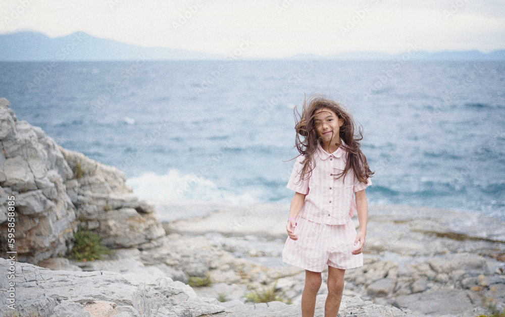 Beautiful young girl in a pink suit poses on a rock high above the sea during sunset. Dark clouds all over the sky. Wind in the hair. Aded noise Beautiful young girl in a pink suit poses on a rock high above the sea during sunset. Dark clouds all over the sky. Wind in the hair. Aded noise