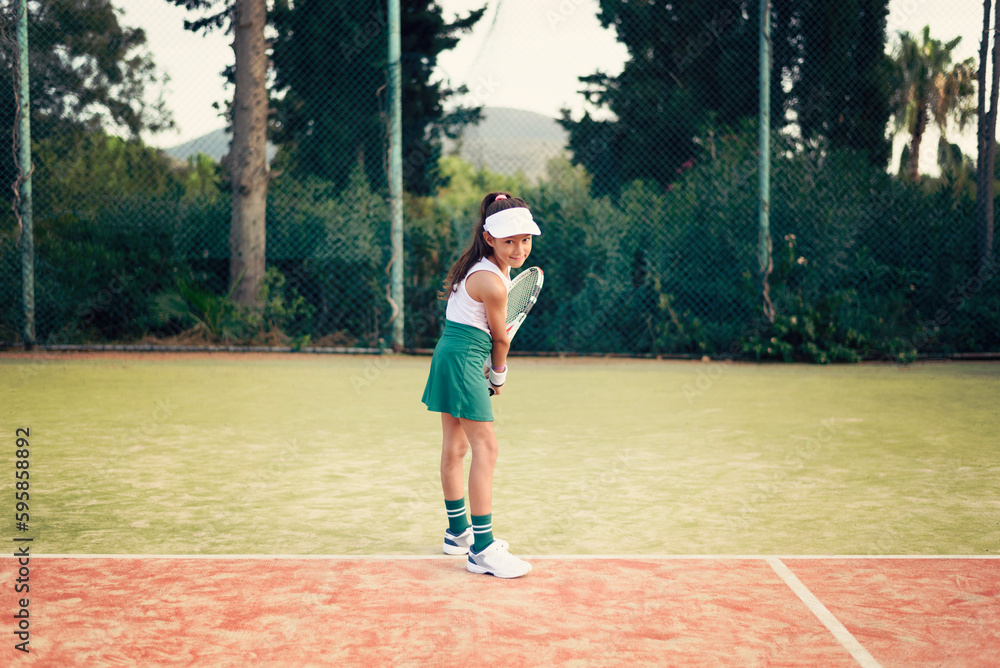 Young girl, tennis player with green skirt, white top and hat, playing tennis on a clay tennis court on a sunny day
