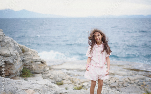 Beautiful young girl in a pink suit poses on a rock high above the sea during sunset. Dark clouds all over the sky. Wind in the hair. Aded noise © sevdastancheva