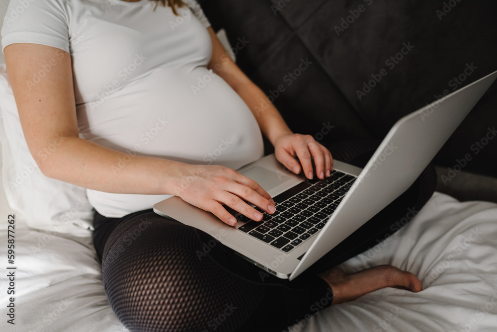 Naklejka premium Pregnant student girl studying at home, learning webinar, workshop. Woman writing notes and using a laptop in bedroom. Female have computer on knees, work as freelancer in bed. Remote work, overwork.