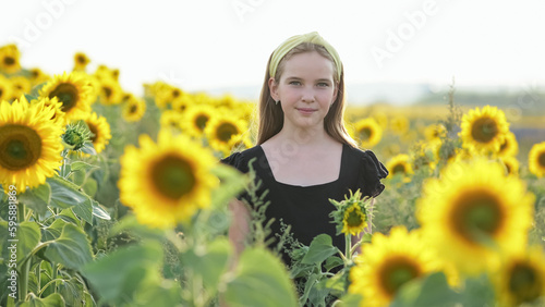 Teen girl stands against blurry blooming sunflower field. Schoolgirl wearing yellow headband looks with amused expression