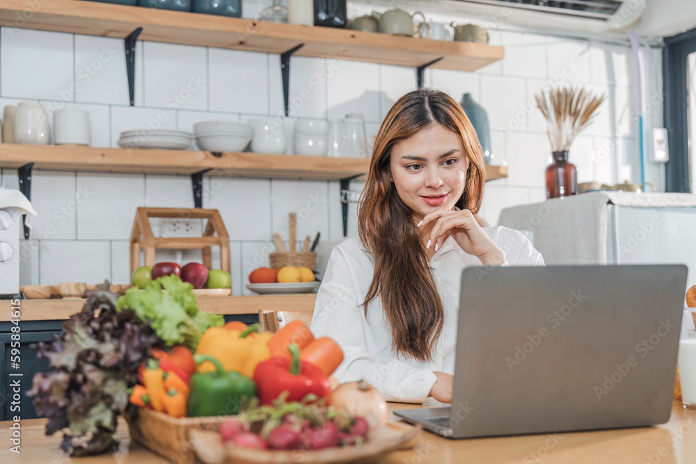 Young happy smiling woman using laptop while cooking vegan food in the modern kitchen, smiling hipster girl preparing healthy vitamin salad with vegetables, eating healthy lifestyle Young happy smiling woman using laptop while cooking vegan food in the modern kitchen, smiling hipster girl preparing healthy vitamin salad with vegetables, eating healthy lifestyle