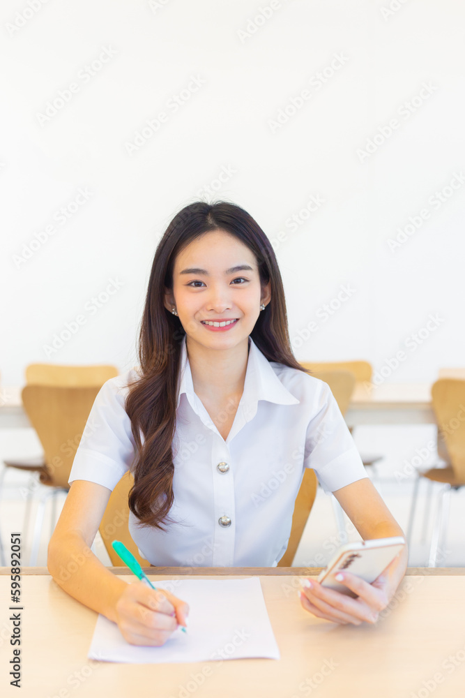 Young Asian woman student in uniform using smartphone and writing something about work. many documents on table her face with smiling in working at to search information for study report at university