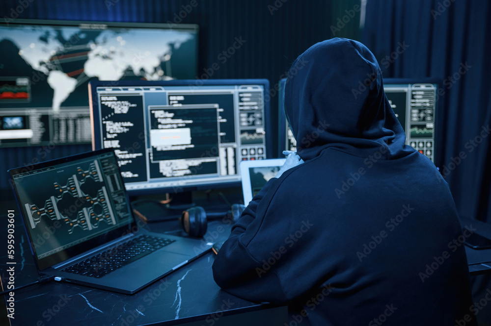 Black colored hoodie. Young professional female hacker is indoors by computer with lot of information on displays Black colored hoodie. Young professional female hacker is indoors by computer with lot of information on displays