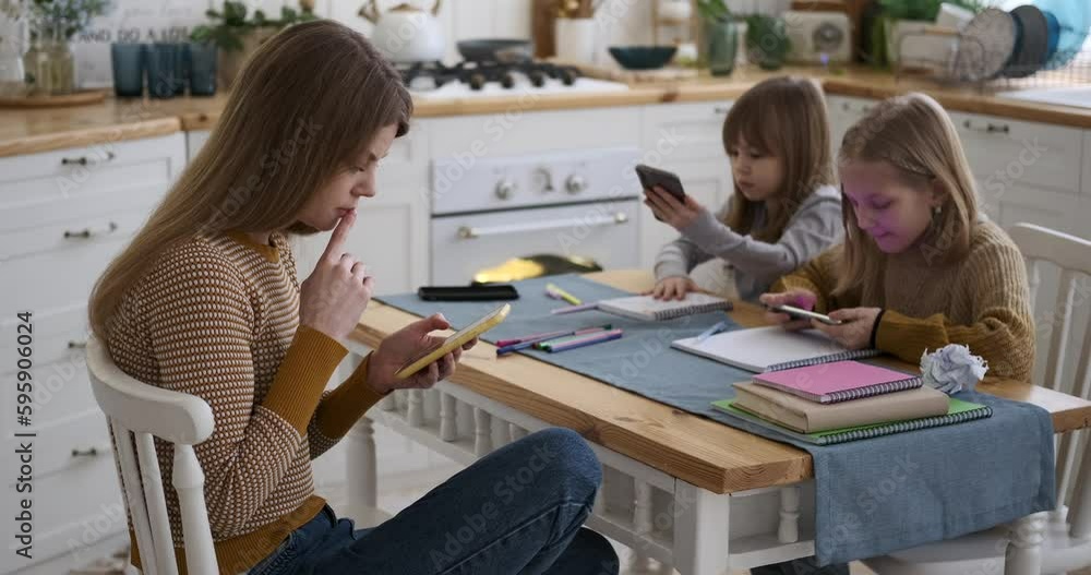 Slow motion shot of young mother scrolling on social media over smartphone and daughters playing games on phones while sitting at table at home Slow motion shot of young mother scrolling on social media over smartphone and daughters playing games on phones while sitting at table at home