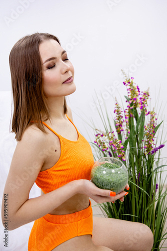 A young girl of European appearance with closed eyes in orange underwear holds in her hands a jar with green granules of wax for depilation. Vertical photo