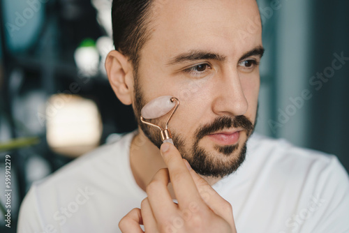Confident man using jade facial massage roller at home interior. Beauty treatment. Facial treatment, cosmetology. Facial skincare.