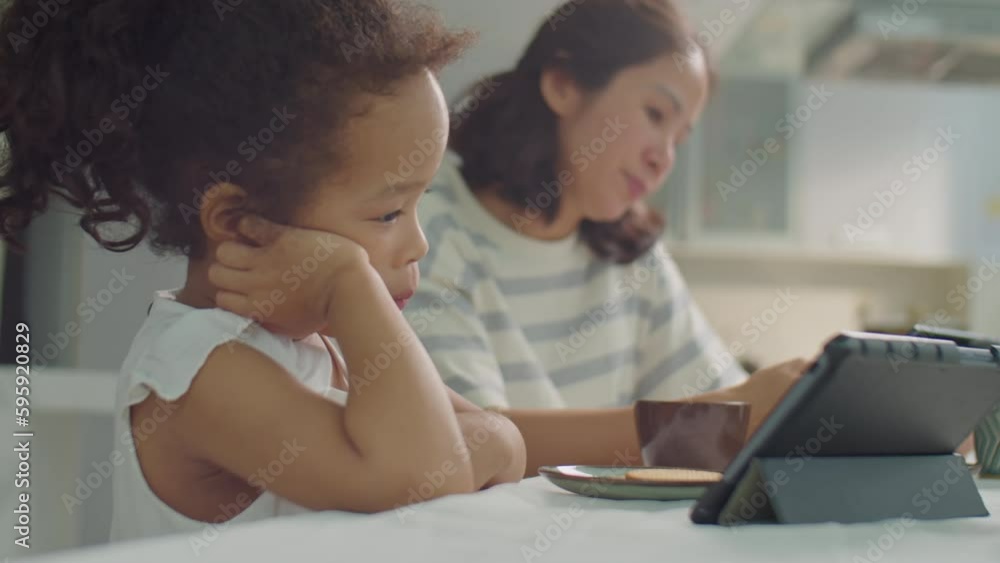 Rack focus shot of Asian mother using smartphone and little daughter watching something on tablet while sitting together at kitchen table during meal Rack focus shot of Asian mother using smartphone and little daughter watching something on tablet while sitting together at kitchen table during meal