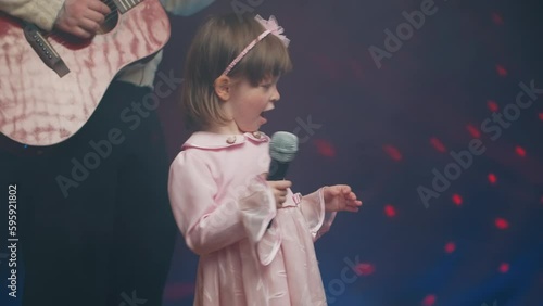 Little girl on stage in vintage dress, she sings into microphone and dances, her father plays acoustic guitar. Color music is shining and smoke is billowing. Father and Child Day, performance on stage