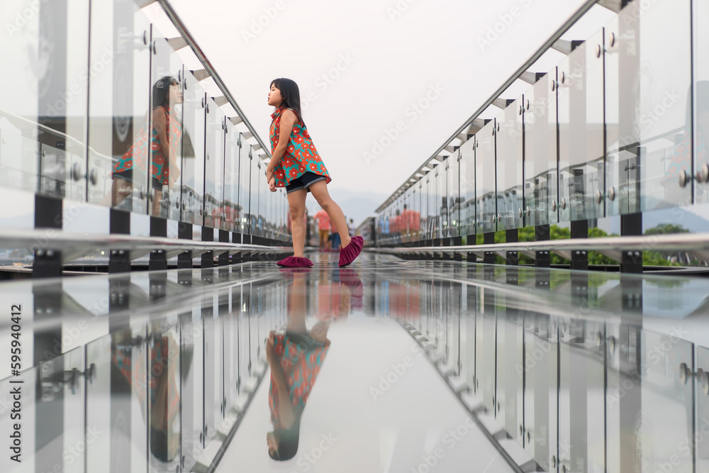 Asian child or kid girl happy standing on skywalk bridge with reflection and clear glass floor on mountain view and fear of heights for vacation travel and holiday relax at Kanchanaburi in Thailand