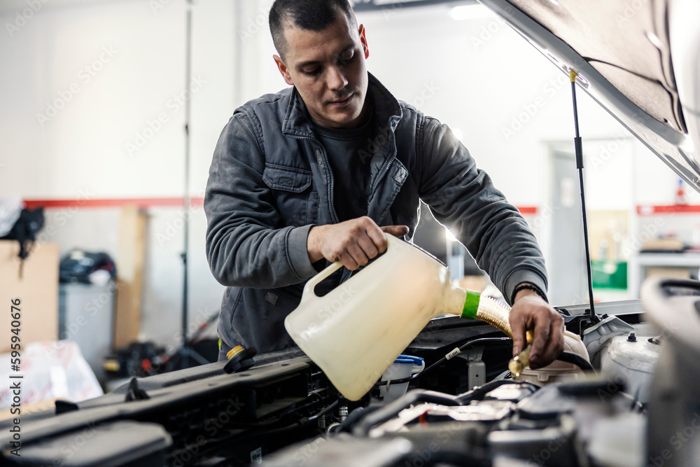 An auto mechanic is pouring oil into the car engine under the hood while standing at garage. An auto mechanic is pouring oil into the car engine under the hood while standing at garage.