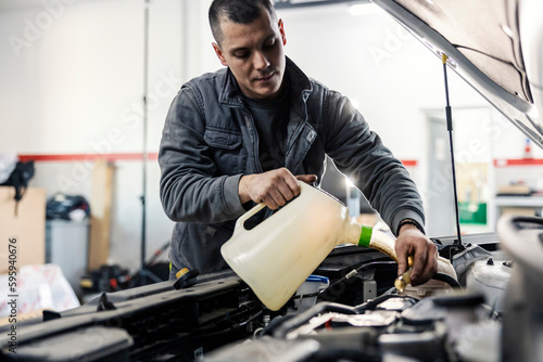 An auto mechanic is pouring oil into the car engine under the hood while standing at garage.