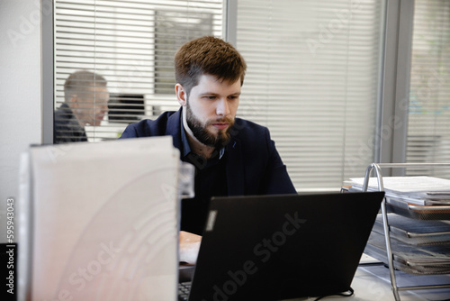 A young man with a beard in a dark blue jacket sits in the office in front of a laptop