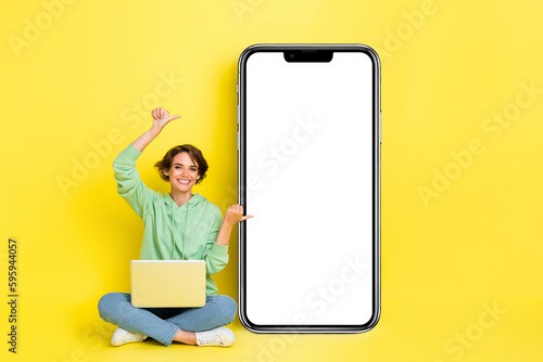 Full length photo of cheerful sweet girl dressed green sweatshirt pointing thumbs modern device empty space isolated yellow color background