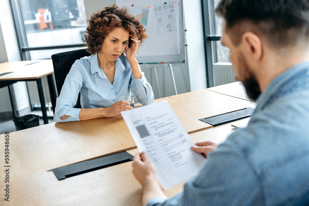 Fototapeta premium Recruitment concept. African American woman is being interviewed for a new job in the office, stressed, anxious, worried, hiring manager studies her resume, asks questions