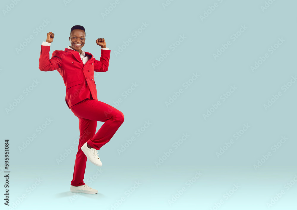 Happy emotional stylish african american man in suit sincerely rejoices in his success isolated on pastel light blue background. Joyful man in red suit happily clenching his fists near copy space.