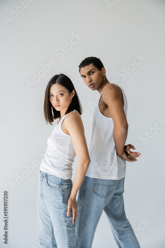 young and trendy interracial couple in white tank tops and jeans looking at camera isolated on grey.