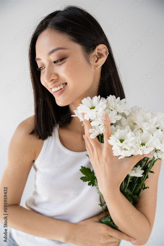 brunette asian woman with cuff earring smiling near white flowers isolated on grey.