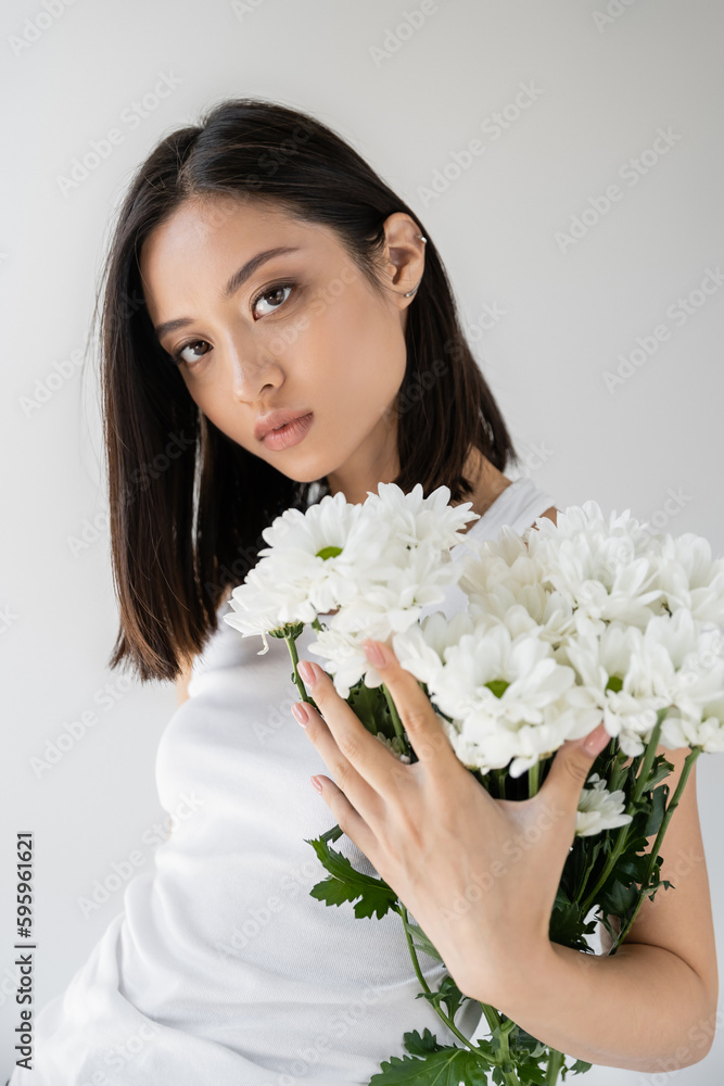 brunette asian woman with perfect skin and natural makeup looking at camera near white flowers isolated on grey. brunette asian woman with perfect skin and natural makeup looking at camera near white flowers isolated on grey.