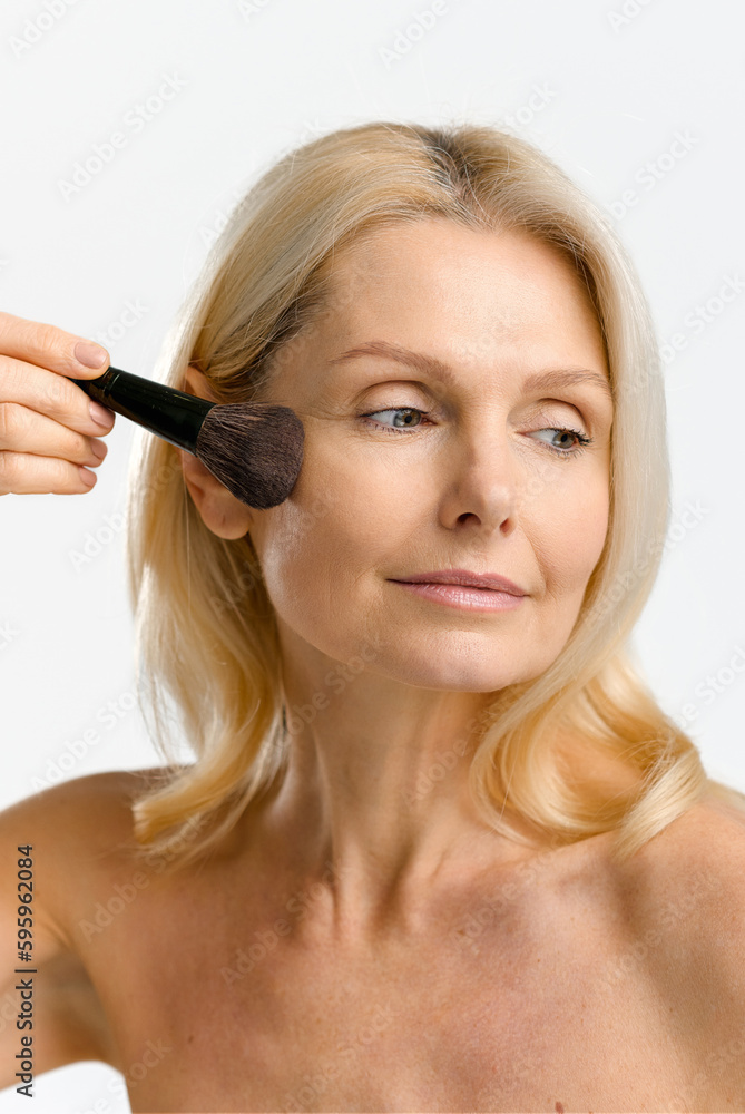 Vertical portrait of charming blonde 40s 50s middle aged woman applying makeup with brush isolated on white