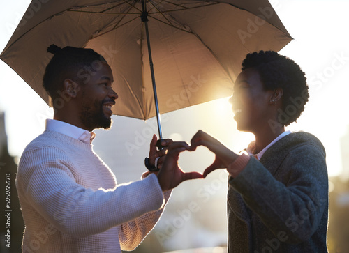 Their love blooms throughout all seasons. a young man and woman making a heart shape with their hands while standing under an umbrella together on a rainy day in the city. © K Davis/peopleimages.com