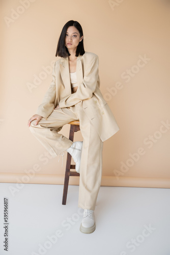 Fotografie brunette asian woman in stylish pantsuit and boots sitting on stool on beige background