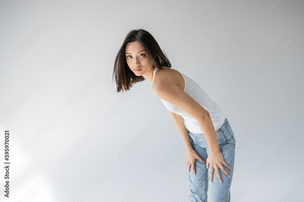 young and slender asian woman in jeans and tank top looking at camera on grey background. young and slender asian woman in jeans and tank top looking at camera on grey background.