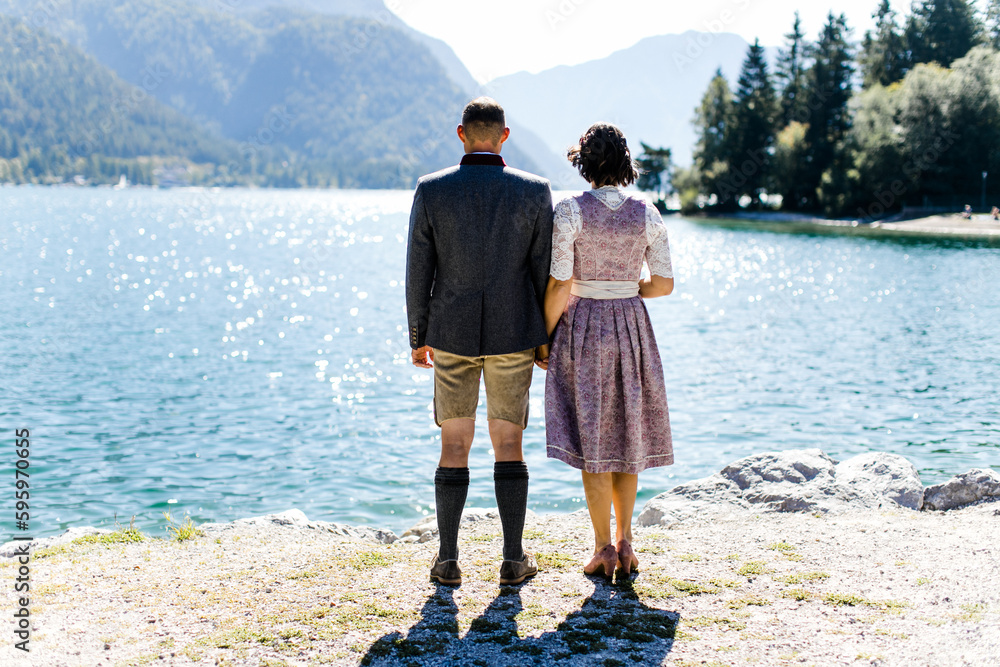 Trachtenpaar In Dirndl Und Lederhose Von Hinten Am See In Tirol Stock trachtenpaar-in-dirndl-und-lederhose-von-hinten-am-see-in-tirol-stock
