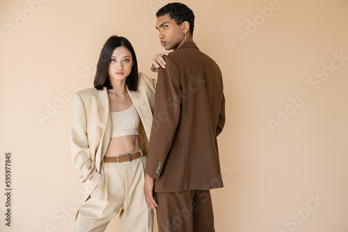 asian woman in trendy pantsuit looking at camera while posing with hand in pocket near african american man isolated on beige.