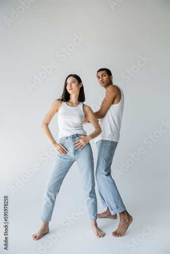 full length of stylish asian woman posing with thumbs in pockets of jeans near barefoot african american man on grey background.