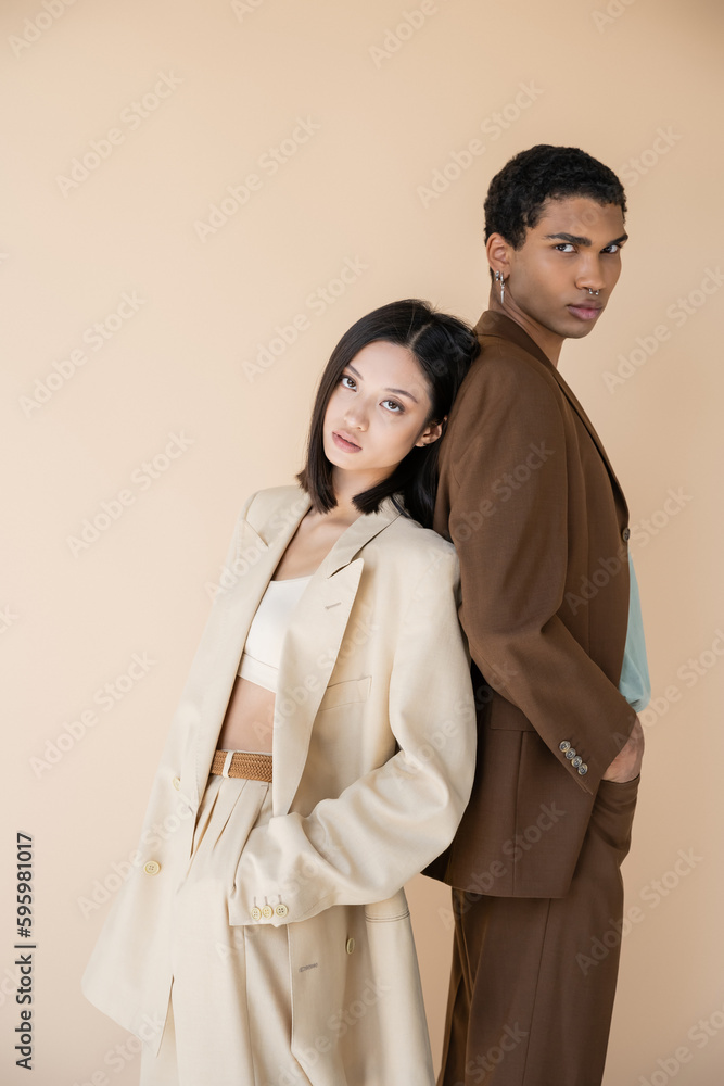 trendy interracial couple in suits standing back to back with hands in pockets isolated on beige. trendy interracial couple in suits standing back to back with hands in pockets isolated on beige.