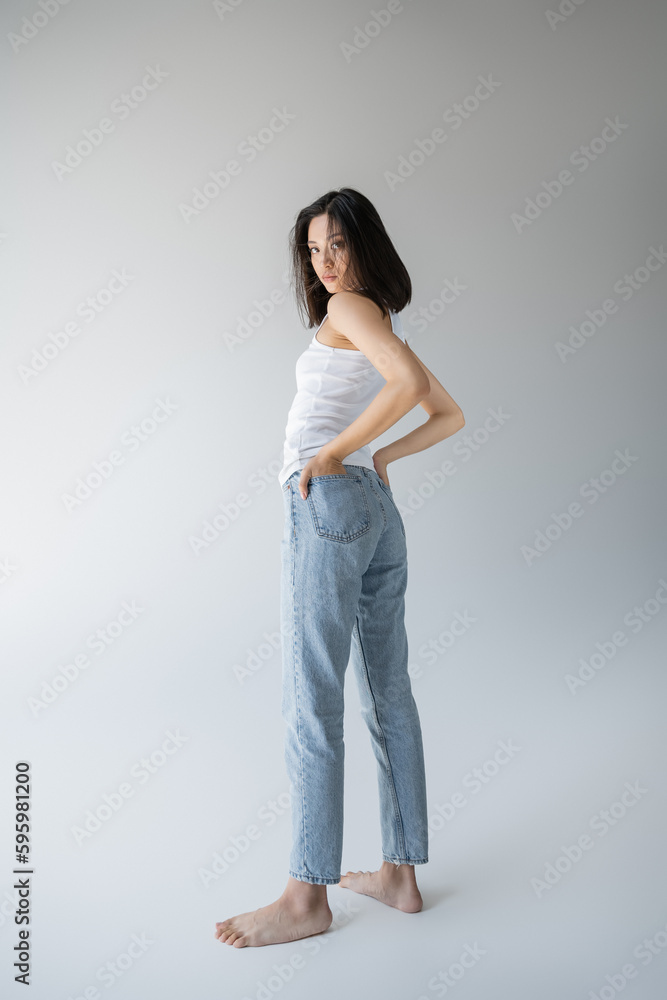 full length of barefoot asian woman in white tank top and blue jeans holding hands in back pockets on grey background. full length of barefoot asian woman in white tank top and blue jeans holding hands in back pockets on grey background.