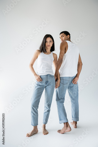 full length of barefoot asian woman posing with thumb in pocket of blue jeans near african american guy on grey background.