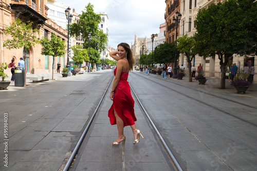 beautiful young woman in a red silk dress stands between the tram tracks on an avenue in seville. The model poses for the photo shoot. Fashion and beauty concept.